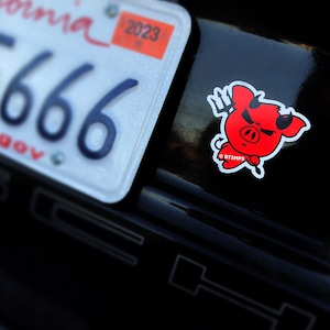 May include: A red cartoon pig with devil horns and a pitchfork, wearing a mischievous expression. The sticker is on a black car with a California license plate that reads "666".