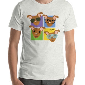 Op de afbeelding: Een lichtgrijs T-shirt met een afbeelding van vier bruine honden die verschillende zonnebrillen dragen. De honden bevinden zich in kleurrijke vierkanten met de tekst "#12MPS" in het midden.