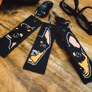 May include: Three black keychains with a white outline of a Doberman Pinscher dog's face. The dog's face is outlined in yellow and orange. The keychains are on a wooden surface. The text "© BT2MPS" is on the bottom right keychain.