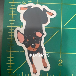 Puede incluir: Una pegatina de perro de dibujos animados negro y marr&oacute;n con orejas rosas. El perro est&aacute; boca abajo y tiene un fondo blanco. La pegatina tiene el texto "&copy;BT2MPS".