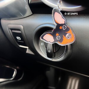 May include: A black and orange dog charm hanging from a car's dashboard. The charm is made of clear acrylic and features a cartoon dog with big eyes. The charm is hanging from a silver chain.
