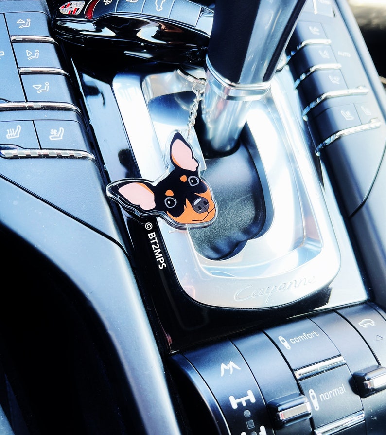 May include: A black and brown dog charm hangs from the gear shift of a black car. The charm is a clear acrylic cutout of a dog's head with the text "BT2MPS" printed on the bottom. The car's gear shift has a silver trim and the word "Cayenne" is printed on the side.