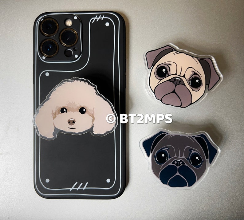 May include: A black phone case with a white border and a clear phone grip with a white poodle design. There are two pug phone grips, one with a light brown and white pug and the other with a black pug.