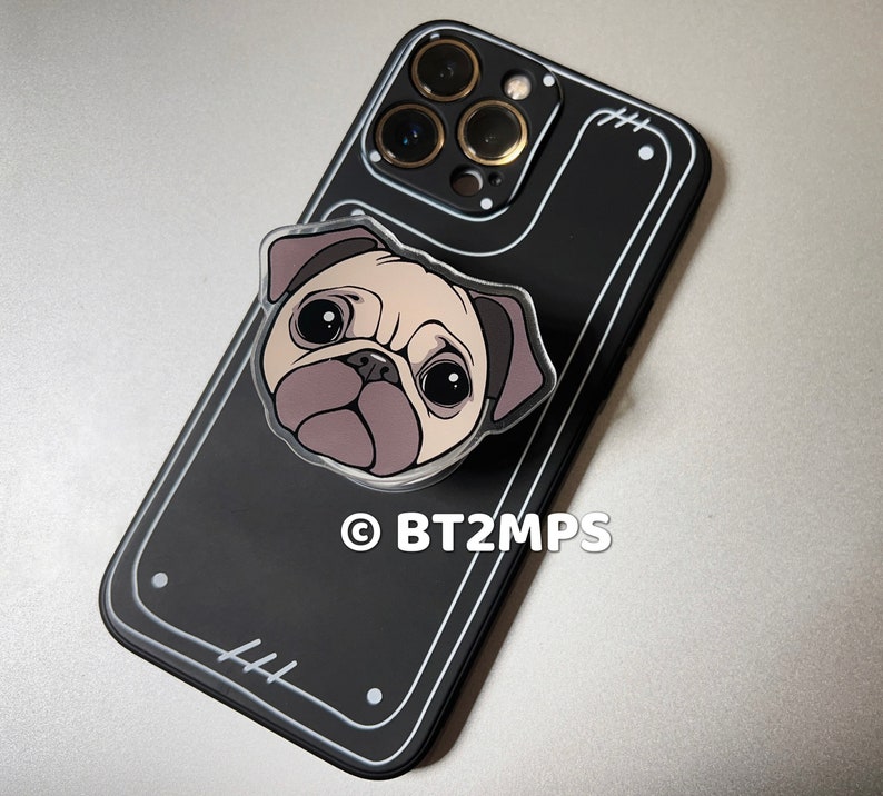 May include: Black phone case with a clear plastic pug phone grip. The phone case has a camera bump with three lenses. The phone grip has a white outline and the text "BT2MPS" is printed on the case.