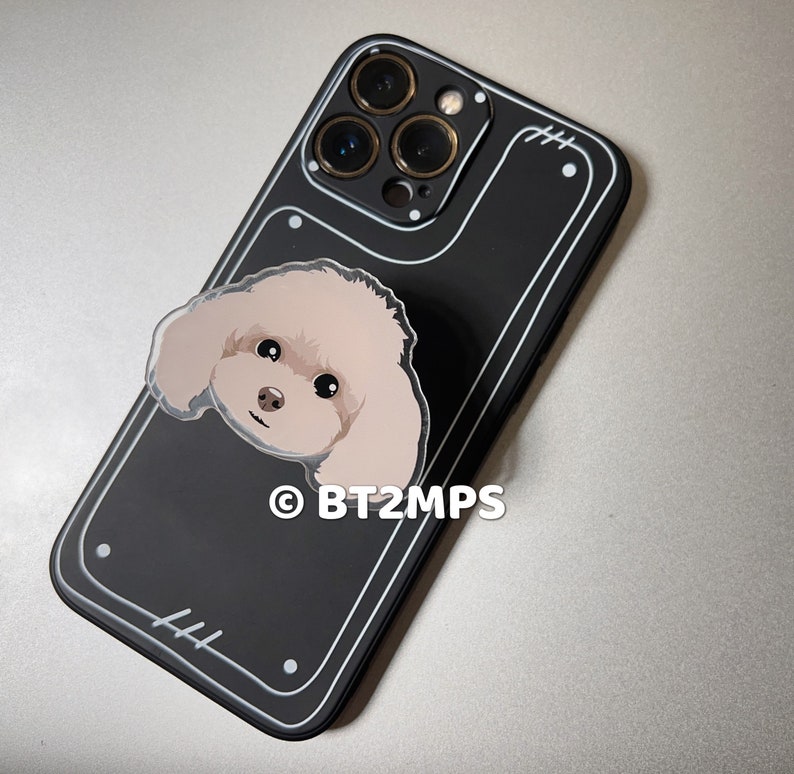 May include: Black phone case with a white border and a small white sticker of a dog's face. The case has a camera lens cutout with four gold rings around it. The text "&copy; BT2MPS" is printed on the case.