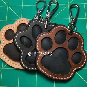 May include: Three leather paw print keychains with silver key rings. The paw prints are brown, black, and tan with white stitching. The keychains are attached to silver key rings.