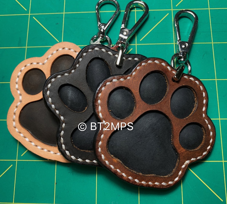 May include: Three leather paw print keychains with silver key rings. The paw prints are brown, black, and tan with white stitching. The keychains are attached to silver key rings.