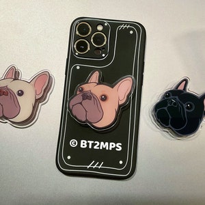 Op de afbeelding: Drie telefoonbeugels met verschillende gekleurde Franse Bulldogs. De beugels zijn gemaakt van helder plastic met een zwarte telefoonhoes op de achtergrond. De telefoonhoes heeft een uitsparing voor de cameralens en de tekst "BT2MPS" erop gedrukt.