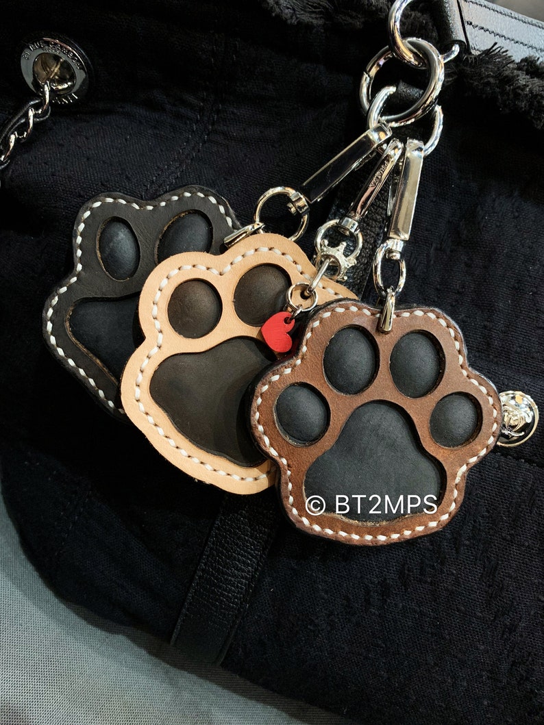 May include: Three leather paw print keychains with white stitching. The keychains are attached to a silver key ring. The paw prints are brown, black, and tan.