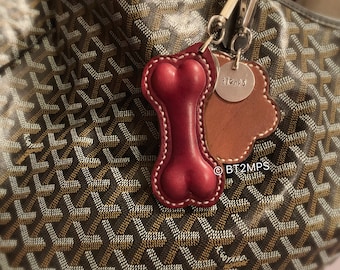 May include: A red leather dog bone keychain with a silver tag that says "BT2MPS" attached to a brown leather loop. The keychain is hanging from a brown and white patterned tote bag.