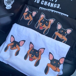 Op de afbeelding: Twee witte T-shirts met een zwart en bruin hondenontwerp. De hond bedekt zijn ogen met zijn poten. De tekst "70 CH4NG3." is gedrukt op een zwart T-shirt eronder.