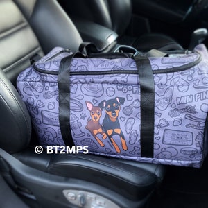Op de afbeelding: Een paarse reistas met zwarte banden en een kleurrijke print met twee dwergpinchers, botten en andere honden gerelateerde items. De tas heeft de tekst "Woof!" en "Min Pins" erop gedrukt.