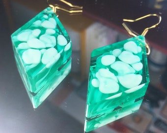 May include: A pair of green and white diamond-shaped earrings with gold hooks. The earrings are made of resin and have a marbled effect.