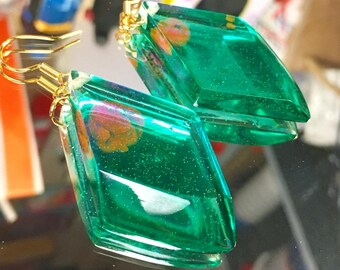 May include: A pair of green diamond-shaped earrings with gold hooks. The earrings are filled with glitter and have a floral design inside.