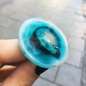 Könnte beinhalten: Ein runder, blauer Glasknopf mit weißem Rand und einem silbernen Ring in der Mitte.