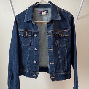 May include: A dark blue denim jacket with a button closure and two front pockets. The jacket has a small logo on the right pocket.