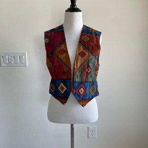 May include: A sleeveless vest with a geometric pattern in shades of red, blue, green, and brown. The vest has a pointed collar and a V-neck.