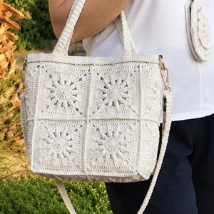 May include: A cream-colored crochet tote bag constructed from square motifs featuring a central sunburst pattern. The bag includes two top handles and a detachable braided shoulder strap with gold-tone metal hardware and a small matching pouch.