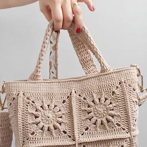 May include: A beige crochet tote bag constructed from four square panels featuring a central sunburst or floral motif. The bag includes two sturdy crochet handles and gold-toned metal hardware attachments for a strap on the sides.