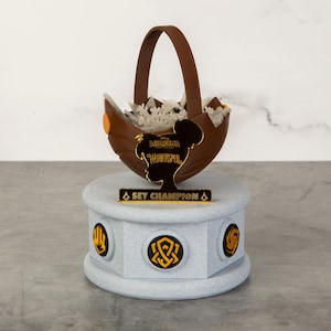 May include: A brown basket-shaped trophy with a handle, filled with white decorative elements, sits atop a gray pedestal. The trophy features a silhouette and the words "LOREANA WARDSPEL" and "SET CHAMPION" in gold lettering. The pedestal has gold symbols.