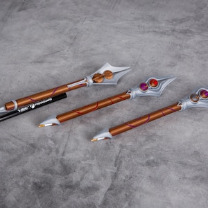 Może przedstawiać: Trzy ozdobne włócznie w kolorze srebrnym i brązowym z czerwonymi i fioletowymi akcentami. Włócznie mają ostry czubek i zaokrąglony uchwyt.