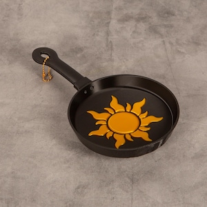 May include: A black frying pan featuring a gold sun emblem with stylised flames in the centre of the cooking surface. The handle includes a circular hole at the end with a small gold-coloured ball chain attached for hanging purposes.