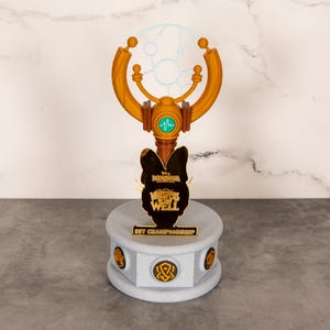 以下が含まれることがあります： 上部に円形デザイン、ベースに「SET CHAMPIONSHIP」の文字が入った金色のトロフィー。 トロフィーにはターコイズ色のアクセントと黒いキャラクターのシルエットがあります。 ベースはグレーで、金のアクセントがあり、高さは約15cmです。