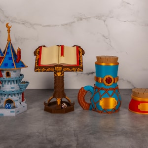 May include: A collection of fantasy-themed tabletop gaming accessories. Includes a multi-tiered castle, a book stand with an open book, a blue mug with a handle, and a red jar, all with decorative details. Dice are visible near the castle and book stand.
