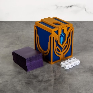 Könnte beinhalten: Eine blaue und goldene Box mit geometrischem Design, ein Stapel lila Karten und sechs weiße Würfel mit schwarzen Zahlen. Die Box hat einen Klappdeckel und einen blauen, juwelenartigen Akzent.