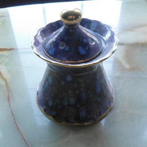 May include: A blue and gold ceramic jar with a lid. The jar has a speckled design and a gold rim. The lid has a gold knob and a small hole in the centre.