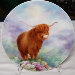 May include: A decorative plate depicting a Highland cow, painted in shades of brown and orange. The cow stands on a rocky outcrop with purple and pink flowers, against a backdrop of a cloudy sky with pastel colours.