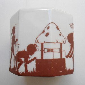 May include: A white ceramic hexagonal box with a brown silhouette design of three figures standing near a well with a mushroom roof. The figures are holding buckets and appear to be fairies.