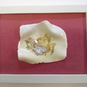 May include: A white ceramic dish with a raised edge, featuring a hand-painted image of a fairy with wings and a blue dress, surrounded by yellow flowers. The dish is framed in a white frame with a pink mount.