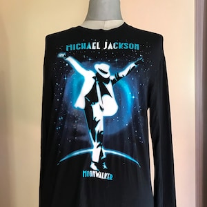 Puede incluir: Camiseta negra de manga larga con un gráfico de Michael Jackson con un traje blanco realizando su movimiento de baile característico sobre un fondo de galaxia azul y negro. El texto "MICHAEL JACKSON" y "MOONWALKER" está impreso en azul en la camiseta.