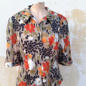 Può includere: Una blusa beige con bottoni e stampa floreale in tonalità arancio, bianco e nero. La blusa ha un colletto e maniche a tre quarti.