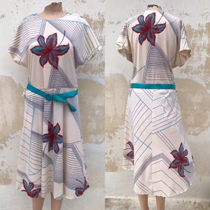 以下が含まれることがあります： 幾何学模様と赤い花柄のアクセントが施された白いドレス。ドレスにはターコイズブルーのベルトと短い袖が付いています。