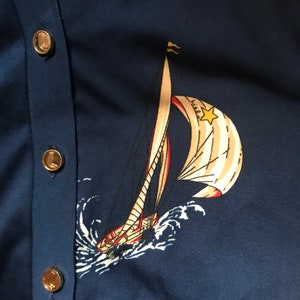 May include: A navy blue cardigan with gold buttons and a white sailboat with red and yellow accents printed on the front. The sailboat is sailing on a blue sea with white waves.