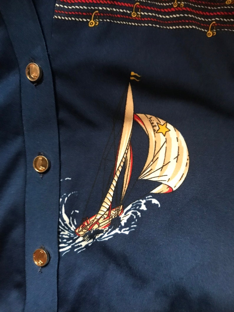 May include: A navy blue cardigan with gold buttons and a white sailboat with red and yellow accents printed on the front. The sailboat is sailing on a blue sea with white waves.