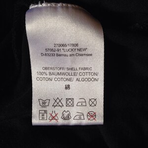 Op de afbeelding: Een wit waslabel met zwarte tekst en symbolen. De tekst luidt "270065/17806 57052-91 "LUCKY NEW" D-83233 Bernau am Chiemsee OBERSTOFF/SHELL FABRIC 100% BAUMWOLLE/COTTON/COTON/COTONE/ ALGOD&Oacute;N 綿". De symbolen geven aan dat het kledingstuk op 30 graden Celsius in de wasmachine gewassen kan worden, in de droger kan worden gedroogd op lage temperatuur, kan worden gestreken op lage temperatuur en niet chemisch gereinigd mag worden.