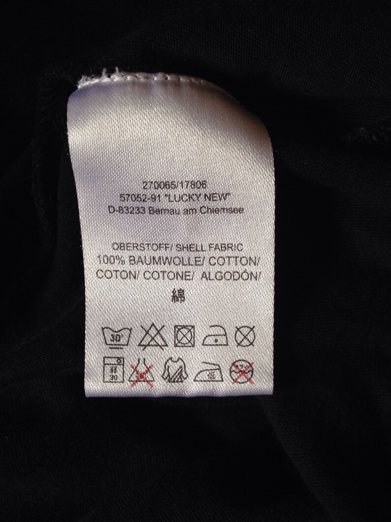 Op de afbeelding: Een wit waslabel met zwarte tekst en symbolen. De tekst luidt "270065/17806 57052-91 "LUCKY NEW" D-83233 Bernau am Chiemsee OBERSTOFF/SHELL FABRIC 100% BAUMWOLLE/COTTON/COTON/COTONE/ ALGOD&Oacute;N 綿". De symbolen geven aan dat het kledingstuk op 30 graden Celsius in de wasmachine gewassen kan worden, in de droger kan worden gedroogd op lage temperatuur, kan worden gestreken op lage temperatuur en niet chemisch gereinigd mag worden.