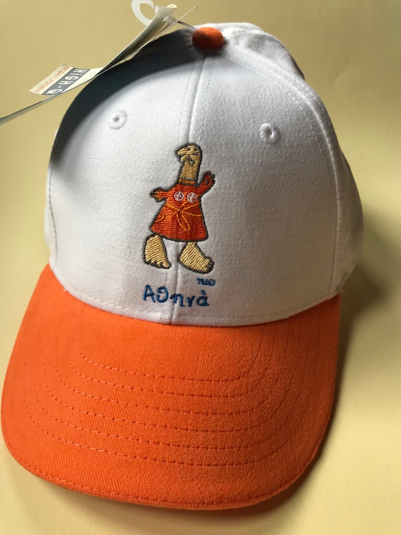 Puede incluir: Gorra de b&eacute;isbol blanca con visera naranja. La gorra presenta un personaje de dibujos animados bordado con un vestido rojo y el texto "the Agnya".