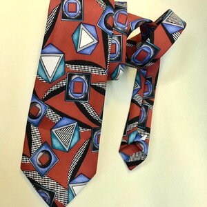 Peut inclure: Une cravate rouge avec un motif géométrique composé de carrés, triangles et cercles bleus, accentué de lignes courbes noires et d'un motif à carreaux noir et blanc. La cravate est nouée, mettant en valeur son design.