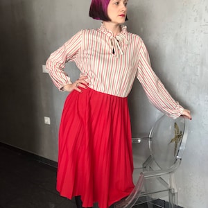 Könnte beinhalten: Eine Frau trägt einen roten Faltenrock und eine gestreifte Bluse mit Schleife. Die Bluse hat rot-grüne Streifen auf cremefarbenem Hintergrund. Sie steht neben einem transparenten Stuhl mit goldenem Design.