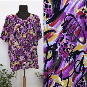 Puede incluir: Una camisa de manga corta con botones de estampado abstracto morado, amarillo y negro. La camisa tiene cuello en V y un corte holgado.