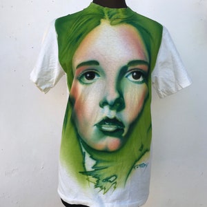 May include: White t-shirt with a green and white airbrushed portrait of a woman's face. The portrait is detailed and realistic, with the woman's eyes looking directly at the viewer. The t-shirt is a vintage style and has a faded look.