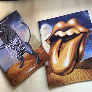 Può includere: Due poster per il concerto dei Rolling Stones Bridges to Babylon World Tour. Il primo poster presenta un leone nero con un cielo blu e nuvole sullo sfondo. Il secondo poster presenta una lingua dorata con un cielo blu e nuvole sullo sfondo. Il testo sul primo poster recita "The Rolling Stones Bridges to Babylon World Tour".
