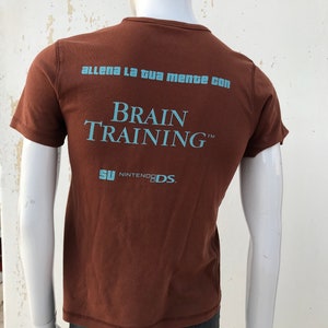May include: Brown t-shirt with the text "Allena la tua mente con Brain Training&trade;  SU NINTENDO DS."