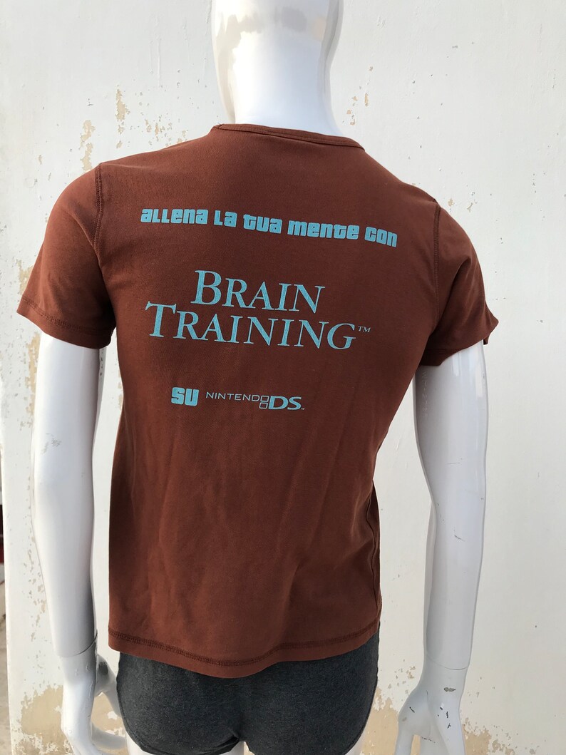 May include: Brown t-shirt with the text "Allena la tua mente con Brain Training&trade;  SU NINTENDO DS."