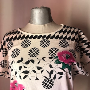 May include: White short-sleeved shirt with a black and white geometric pattern and pink flowers. The shirt has a round neckline and a loose fit.