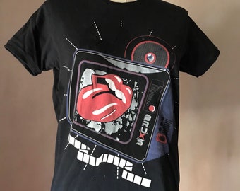 May include: Black t-shirt featuring a graphic of a retro television displaying the Rolling Stones' iconic tongue and lips logo. The design includes a speaker and the band's name. The shirt is displayed on a mannequin.